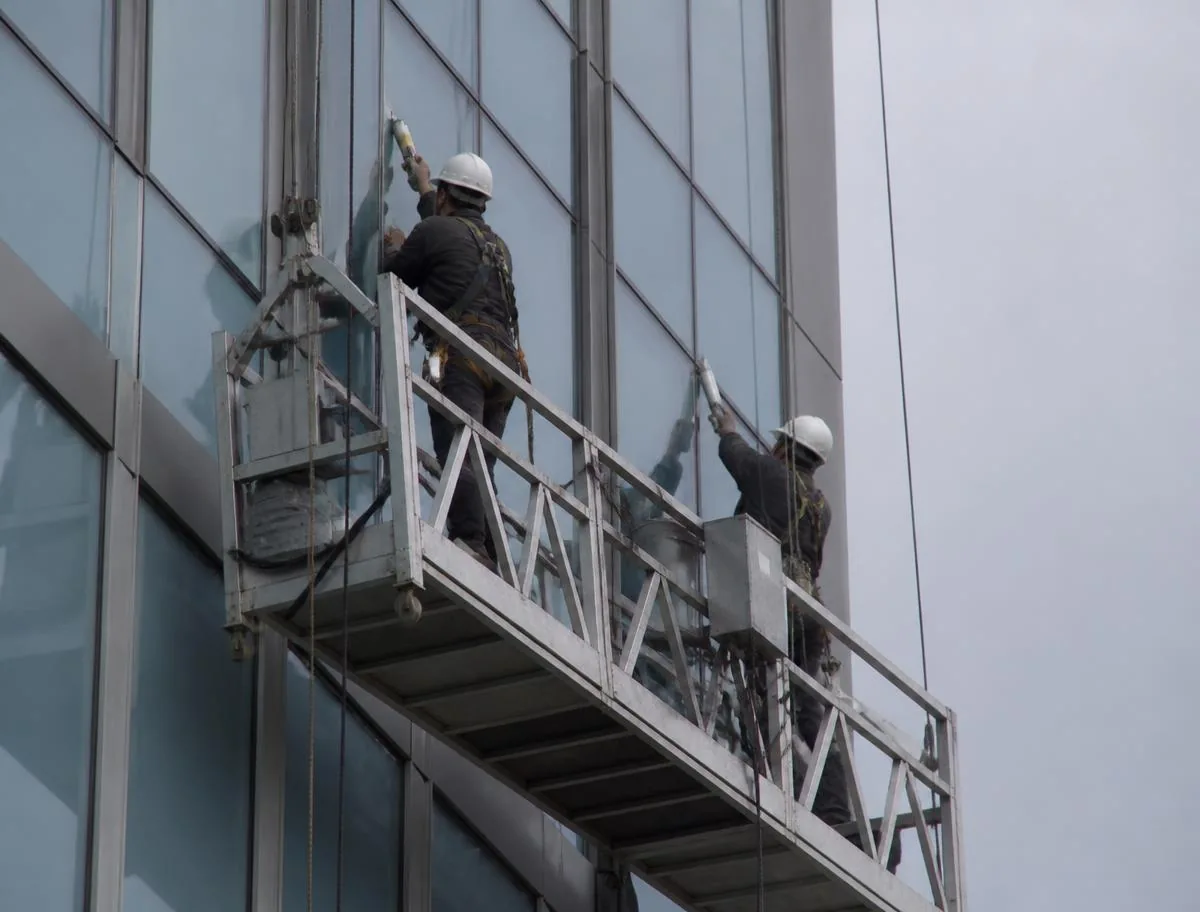 Two workers on a suspended swing stage platform re-caulking vertical joints on a mid-rise glass and aluminum curtain wall